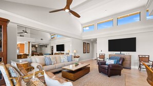 A spacious living room with a high ceiling, a ceiling fan, a large flat-screen TV, and a dining area with wicker furniture.