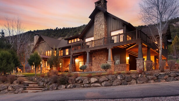 Un'ampia lodge in pietra e legno, su più piani, con un balcone, sullo sfondo di un cielo al tramonto.