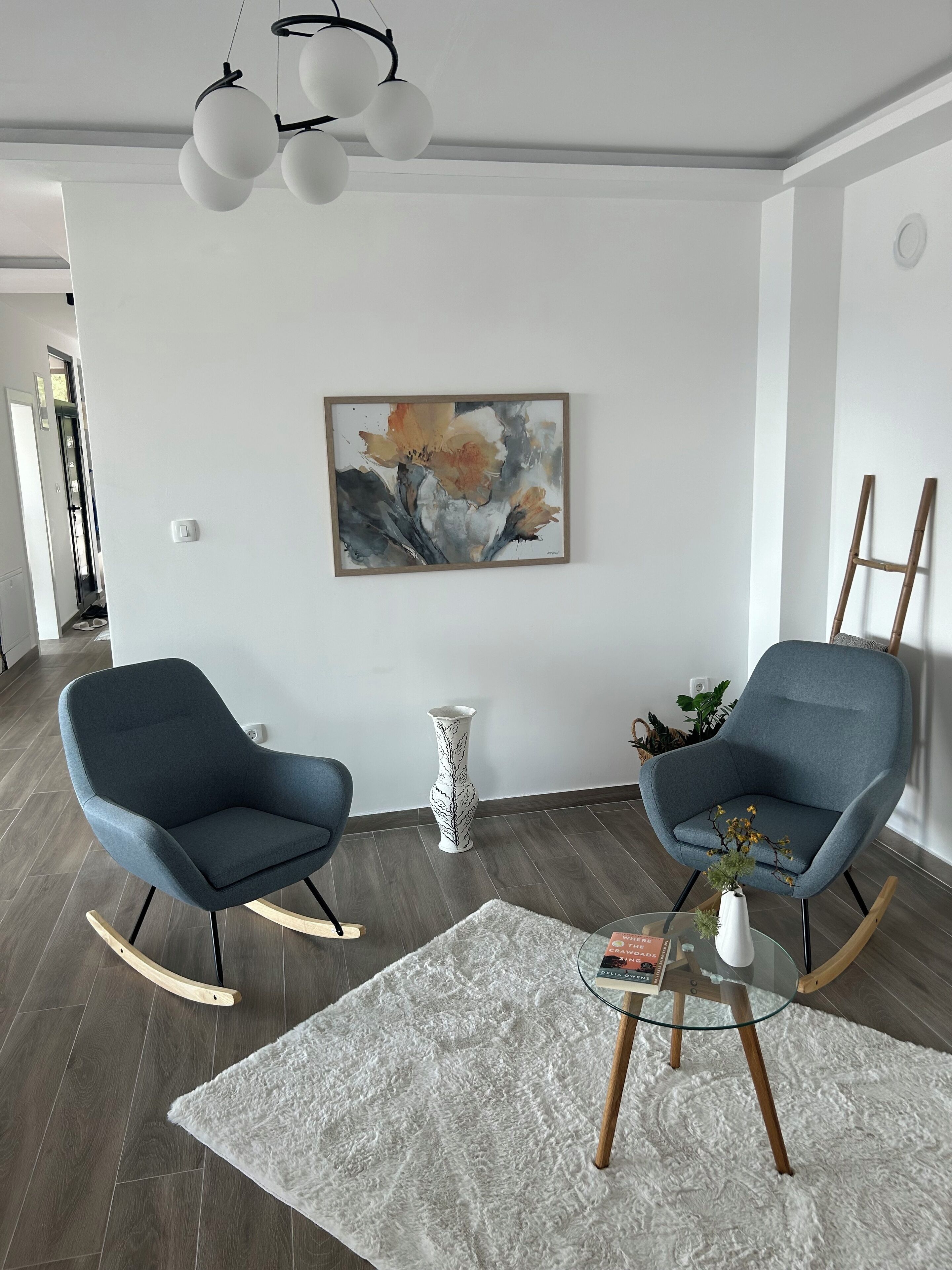Ein modernes Wohnzimmer mit zwei blauen Schaukelstühlen, einer weißen Vase mit Blumen und einem Bild an der Wand.