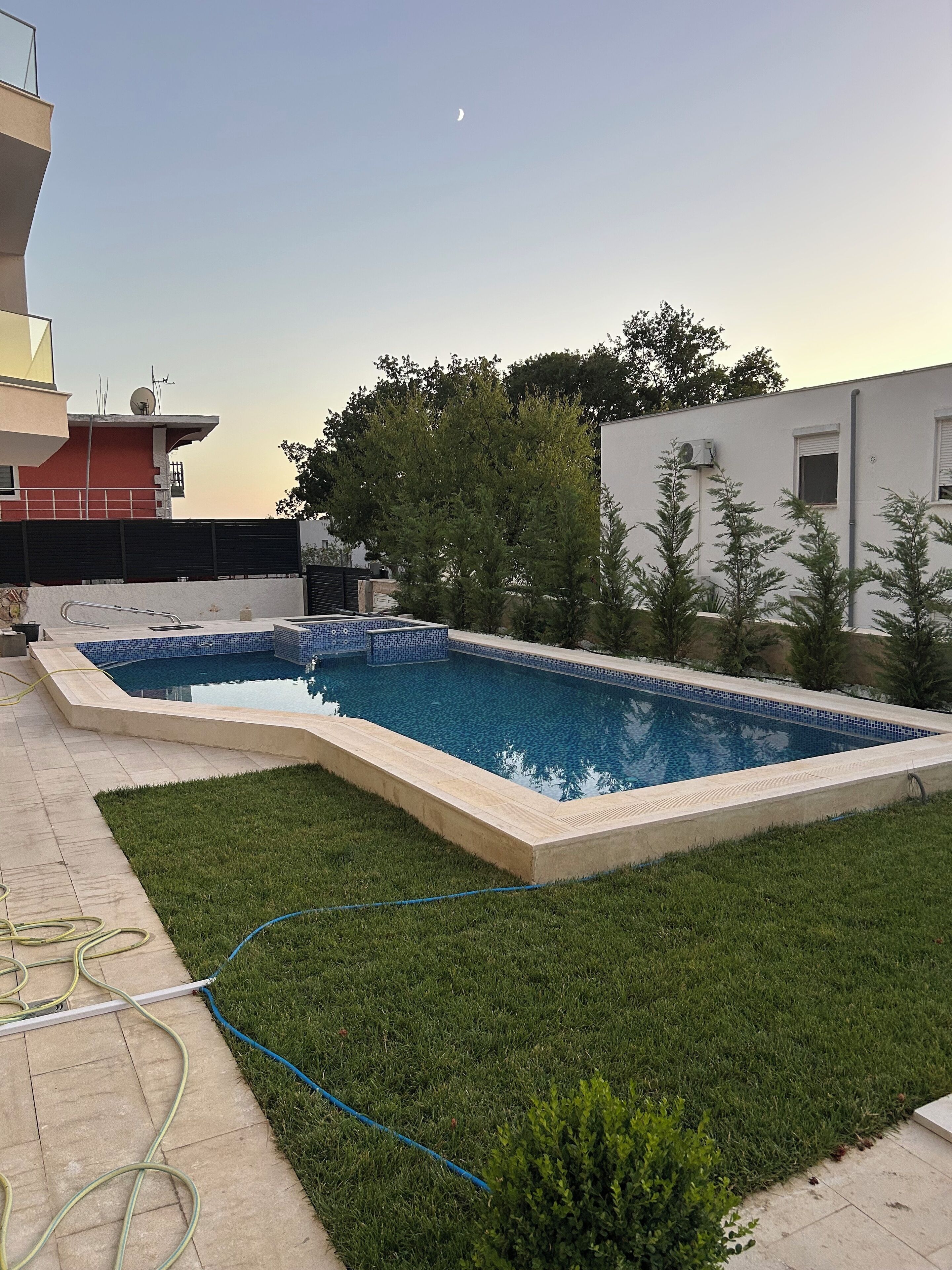 Una piscina rectangular amb vora de rajola, envoltada per un jardí ben cuidat i una manguera visible al terra.