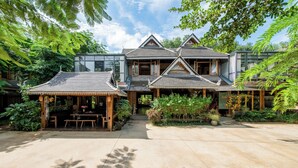 A traditional wooden house with multiple gabled roofs, surrounded by lush greenery and a paved courtyard. - Zhaizili Resort Xishuangbanna  (Xishuangbanna Dai)