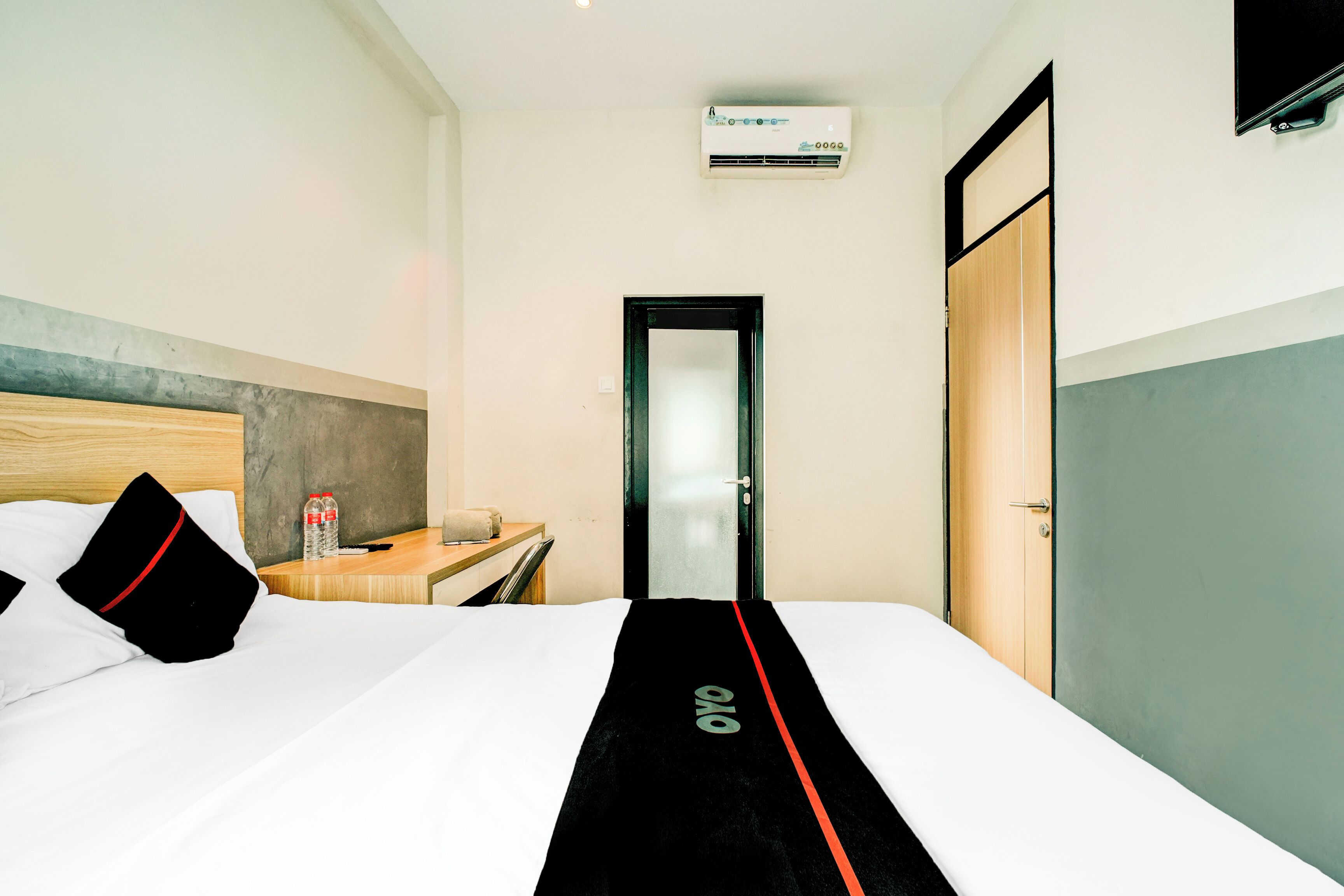 A hotel room with a bed, a wooden headboard, a nightstand with a bottle and remote, a door, and an air conditioning unit.