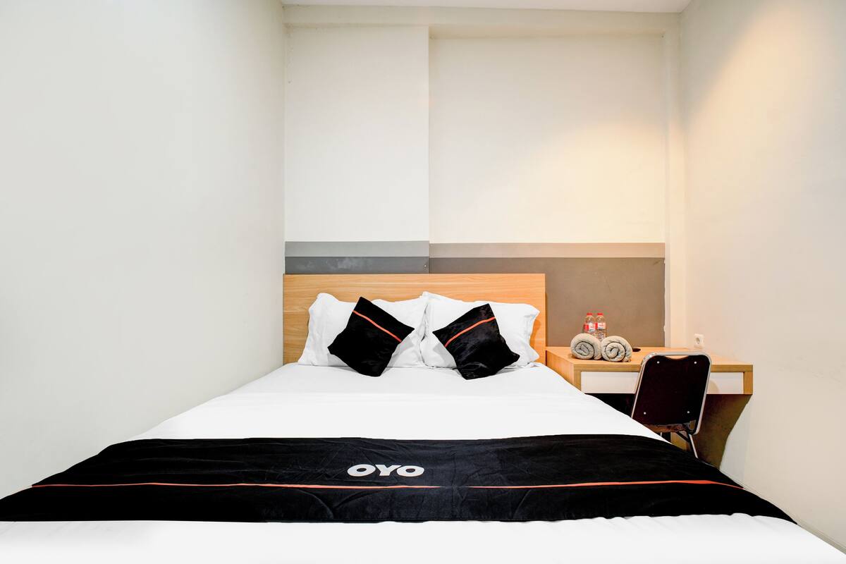 A neatly made bed with a black and white bedspread, two black pillows, and a wooden headboard. A small table with a chair and a bottle of water is visible to the right.