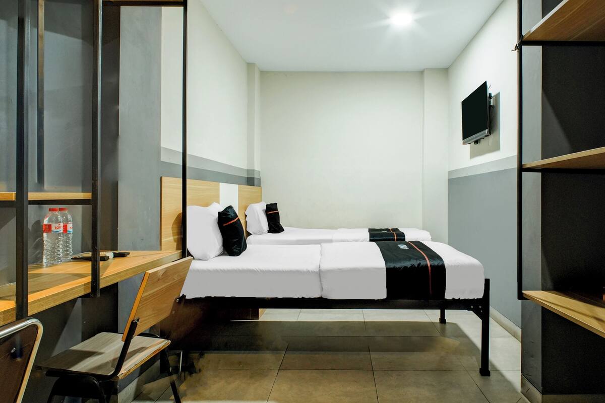 A compact hotel room with a bed, a desk, a chair, a TV, and a bottle of water.
