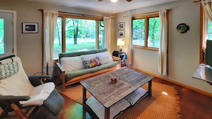 A cozy living room with a sofa, a coffee table, and a lamp. There are windows with curtains, a clock on the wall, and a door.