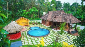 A tropical garden with a swimming pool, a thatched-roof hut, and a brick building with a tiled roof.