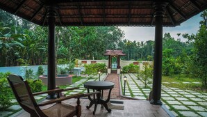 A covered outdoor seating area with a table and chairs, surrounded by a garden with a pathway and a small pavilion in the background.