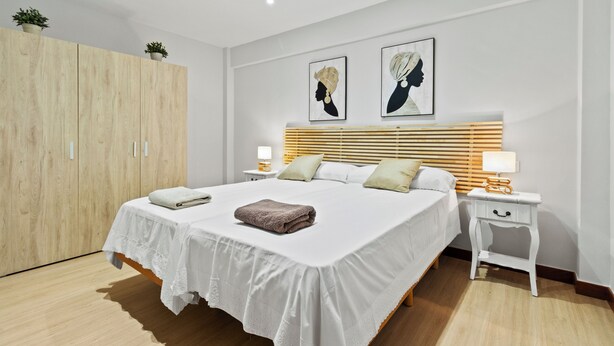 A modern bedroom with a large bed, wooden wardrobe, two bedside tables with lamps, and artwork on the wall.