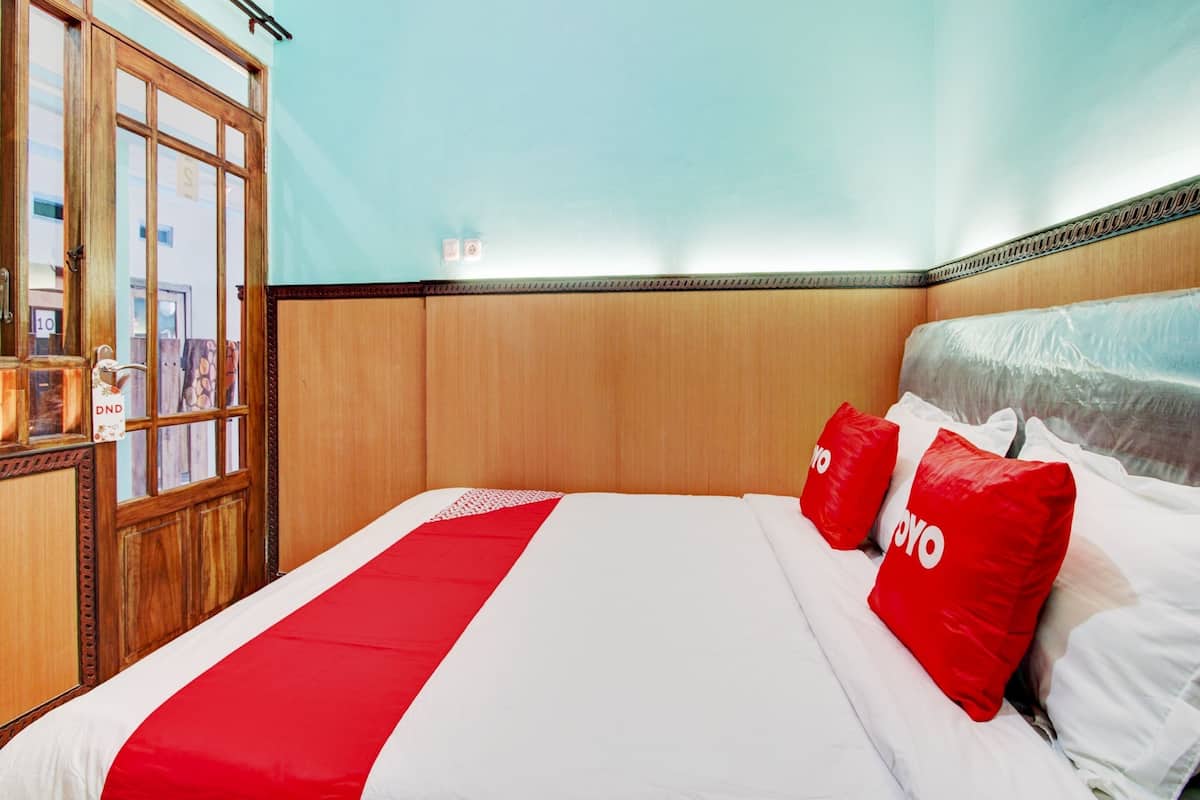 A hotel room with a wooden headboard, a large bed with white and red bedding, and a view of the outside through a glass door.