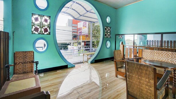 A room with teal walls, wooden flooring, and a large circular window. There are wicker chairs and a small table. - Hotel O Sybilla Cafe & Homestay (Malang)
