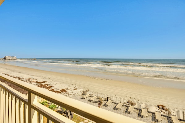 Blick auf einen Strand vom Balkon mit Geländer, ein Steg in der Ferne und ein sandiger Strand mit etwas Vegetation.