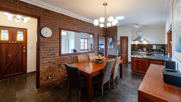 A dining area with a brick wall, wooden dining table, and chairs. There is a clock on the wall and a kitchen area in the background. - Bunbury Bliss - Views and Vibes (East Bunbury)