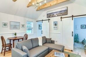 A modern living room with a sectional sofa, a wooden coffee table, and a dining area with a small table and chairs. There are framed pictures on the walls and a chandelier above the sofa. - Osterville Gem Steps to Main St! Relax fireside (Osterville)