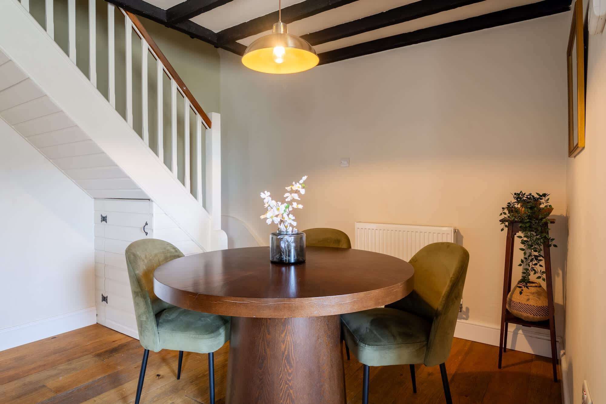 Un coin repas avec une table ronde en bois, deux chaises recouvertes d’un tissu vert, un vase avec des fleurs blanches, une suspension et un escalier avec des rampes blanches.