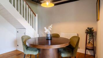 Un coin repas avec une table ronde en bois, deux chaises recouvertes d’un tissu vert, un vase avec des fleurs blanches, une suspension et un escalier avec des rampes blanches.