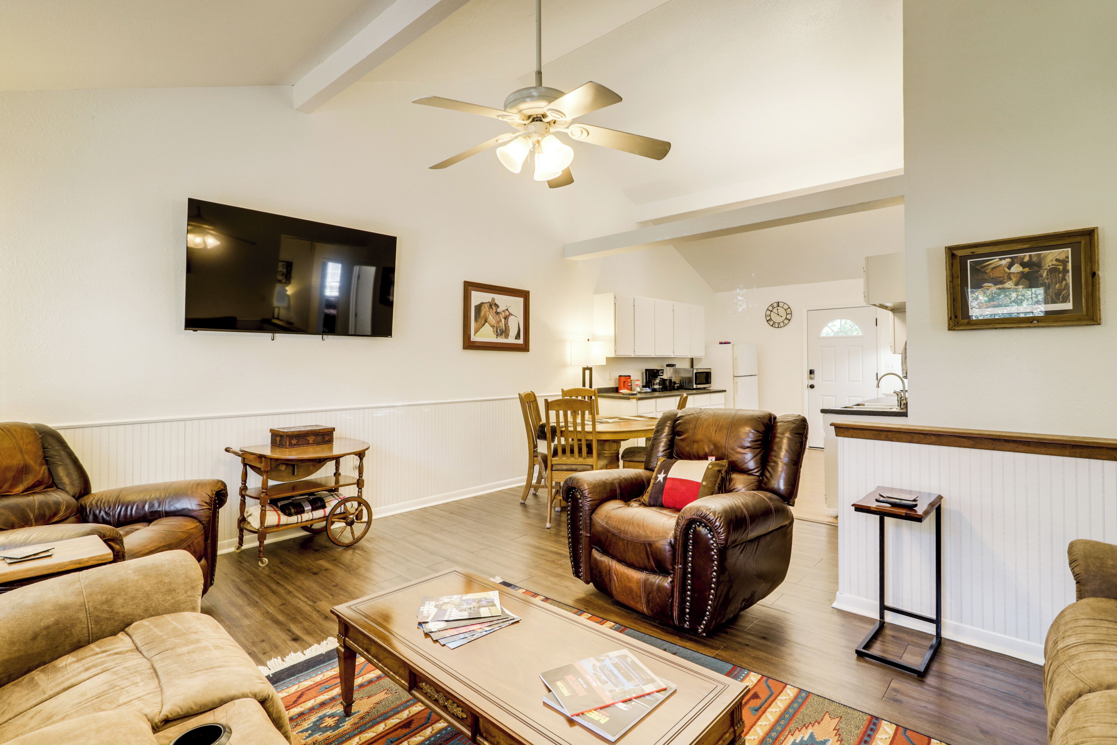 A living room with a sofa, armchair, coffee table, and a dining area with a table and chairs. A television is mounted on the wall, and there is a ceiling fan.