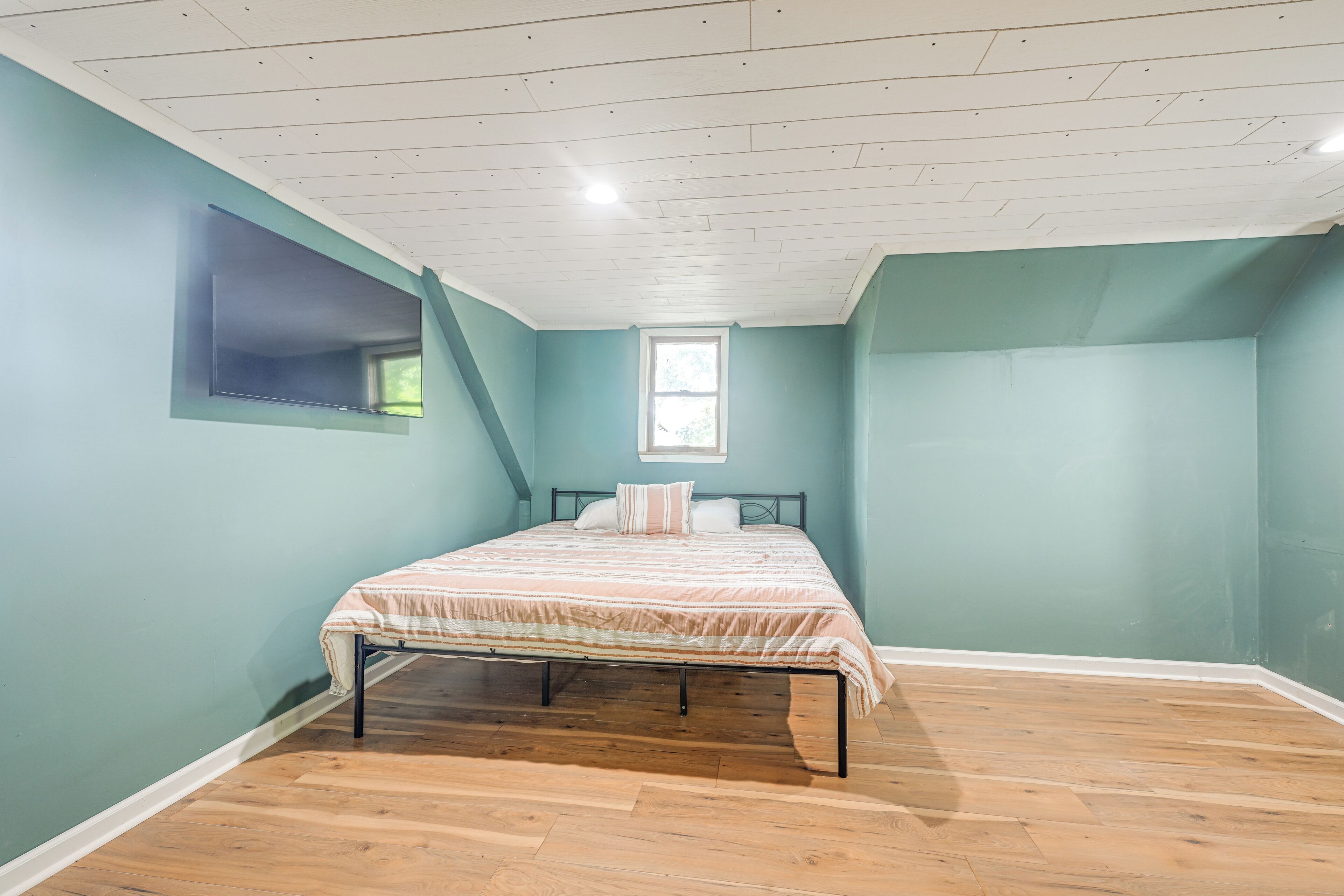 A bedroom with teal walls, a wooden floor, a bed with a striped bedspread, a window, and a ceiling with recessed lighting.