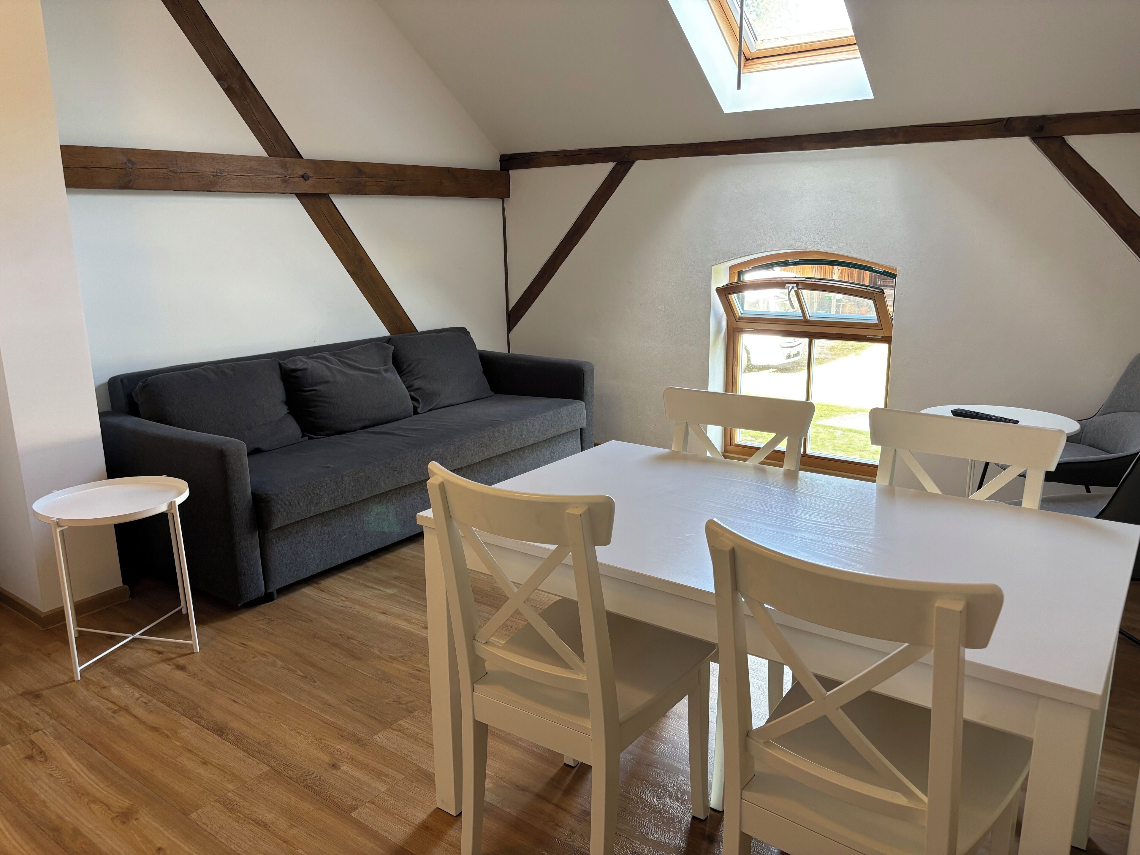 Une pièce avec un canapé gris foncé, une table à manger blanche avec quatre chaises et une petite table ronde. Il y a une lucarne au-dessus du canapé et une fenêtre donnant sur l’extérieur.
