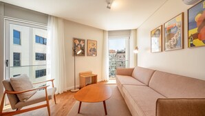 Un salon moderne avec un canapé, un fauteuil et une table basse. Des tableaux encadrés ornent le mur et une vue sur les bâtiments environnants est visible par la fenêtre.
