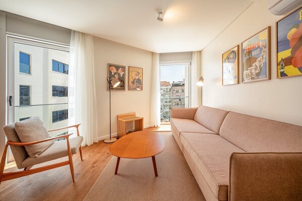 Un salon moderne avec un canapé, un fauteuil et une table basse. Des tableaux encadrés ornent le mur et une vue sur les bâtiments environnants est visible par la fenêtre.