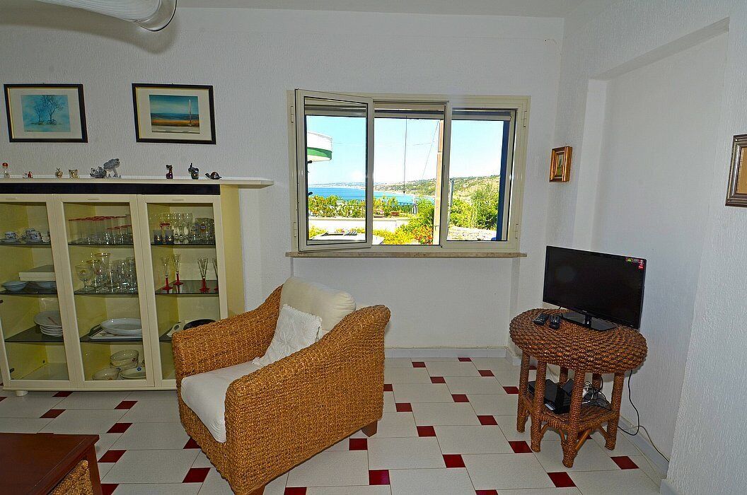 Uma sala de estar com um sofá de vime, uma televisão sobre um suporte trançado, um armário com portas de vidro e uma janela com vista para o mar.