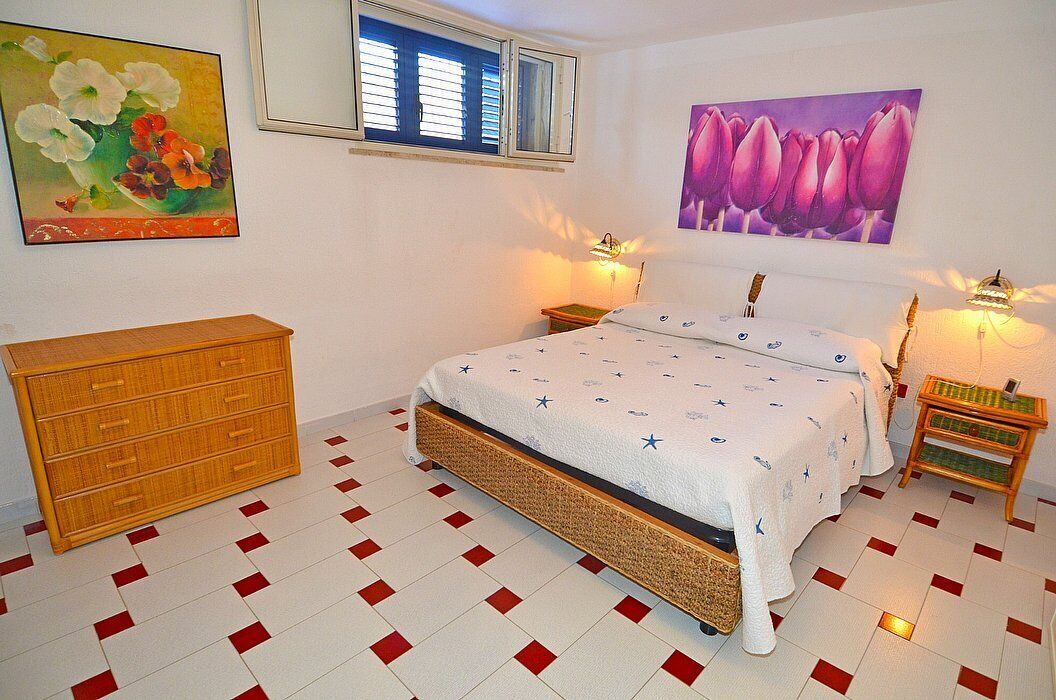 A bedroom with a bed, wooden dresser, two wall-mounted lamps, and two paintings.
