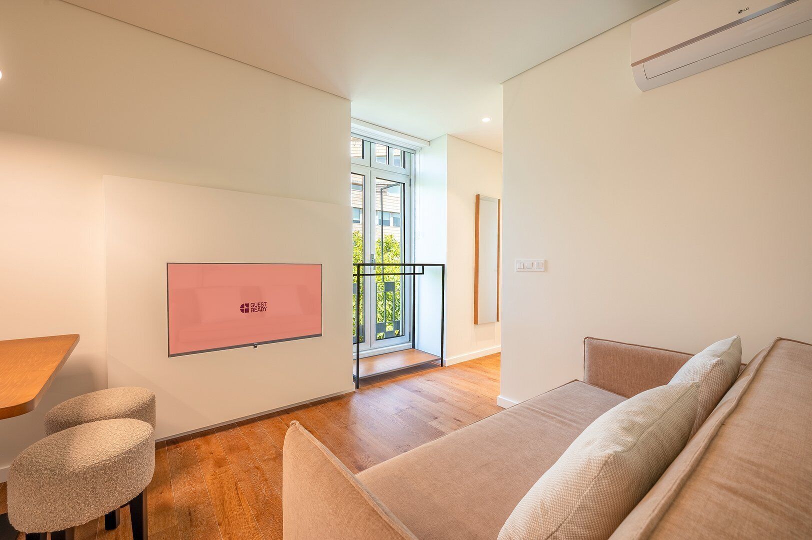 Une pièce moderne et bien éclairée, avec un plancher en bois, un canapé beige, une petite table et un écran rose fixé au mur.