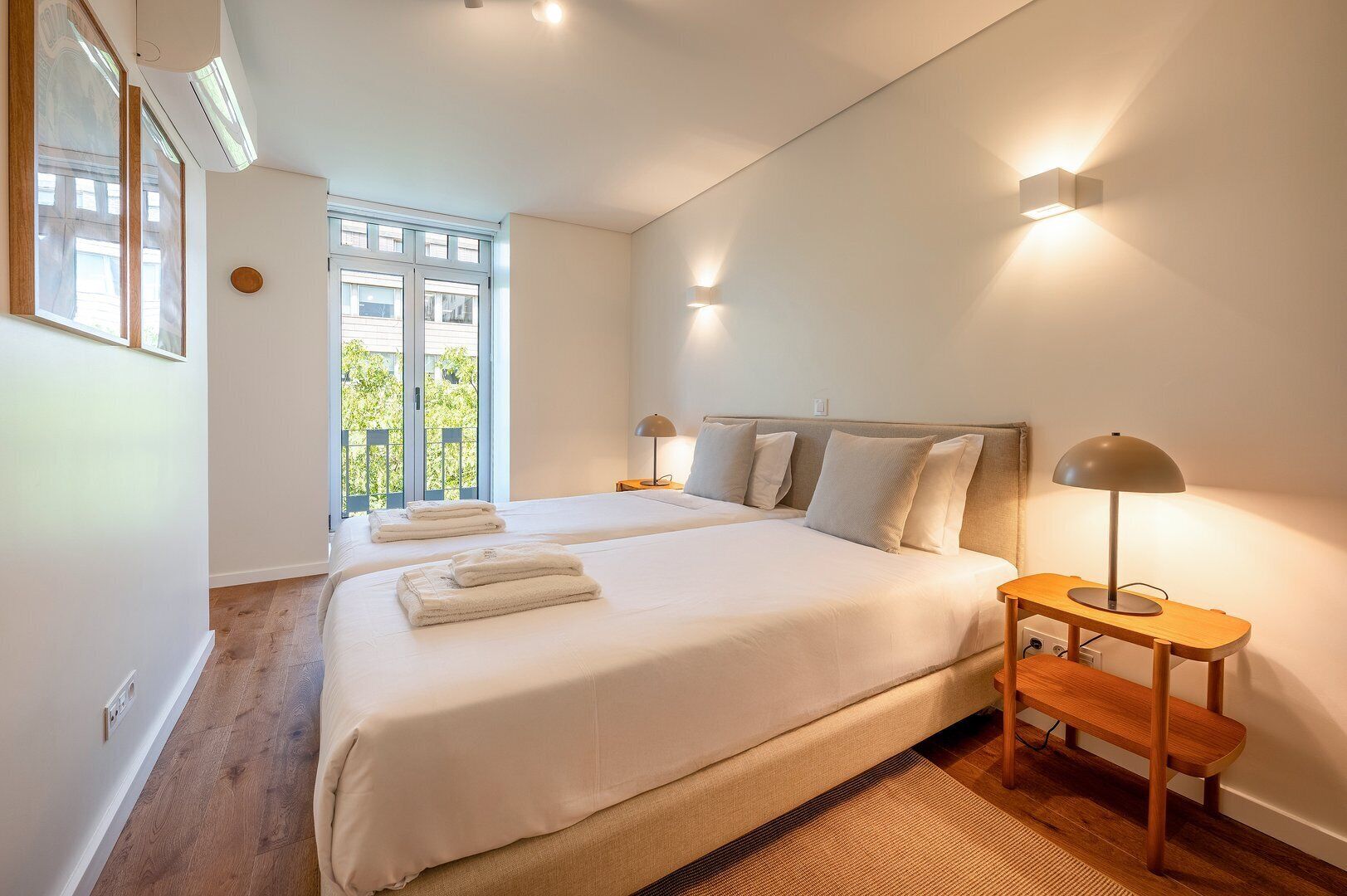A bedroom with two beds, a nightstand, a lamp, and a view of greenery outside.