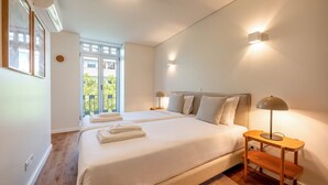 A bedroom with two beds, a nightstand, a lamp, and a view of greenery outside.