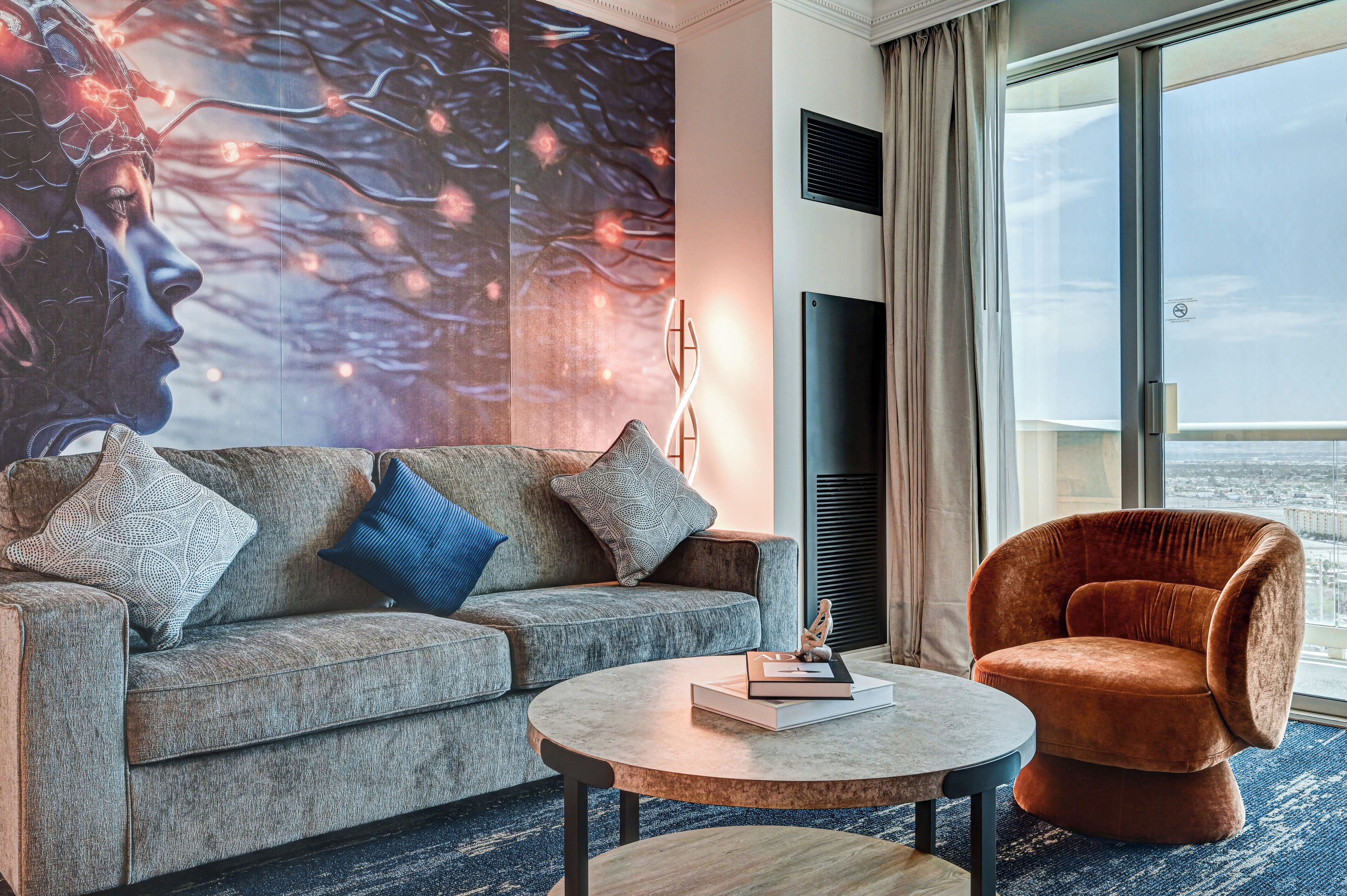 A modern living room with a grey sofa, a round coffee table, and a brown armchair. There is a large wall mural and a window with a view.