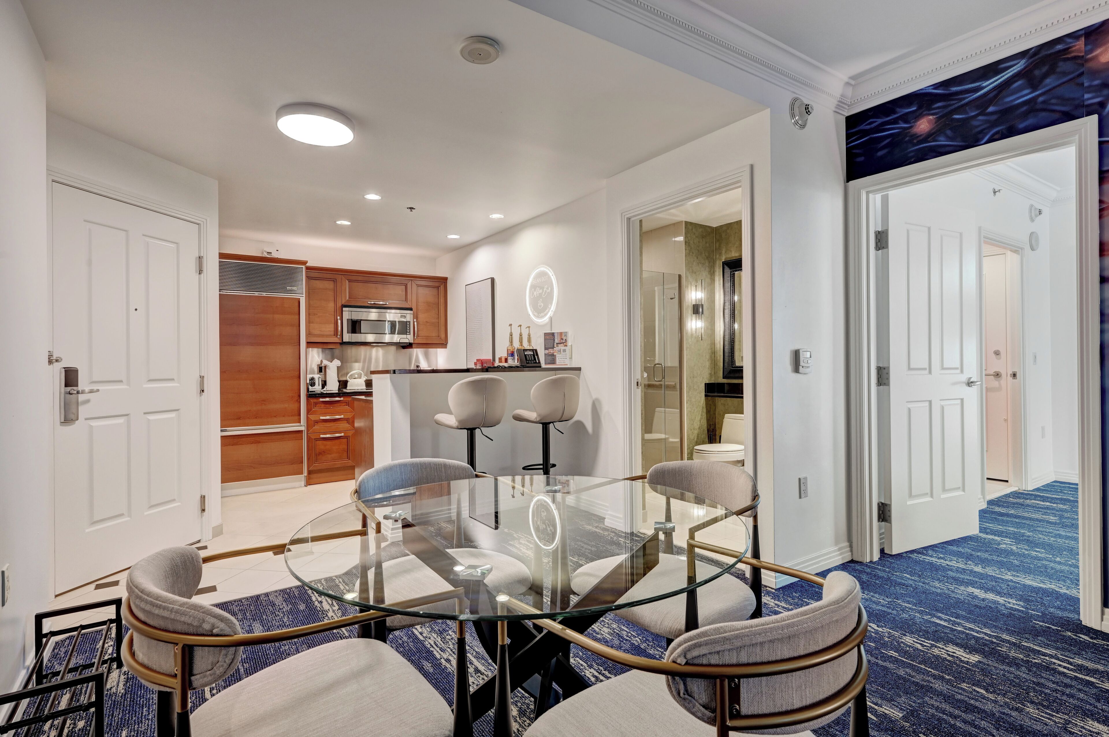 A modern hotel room with a glass dining table, comfortable seating, a kitchenette, and a hallway leading to other rooms.