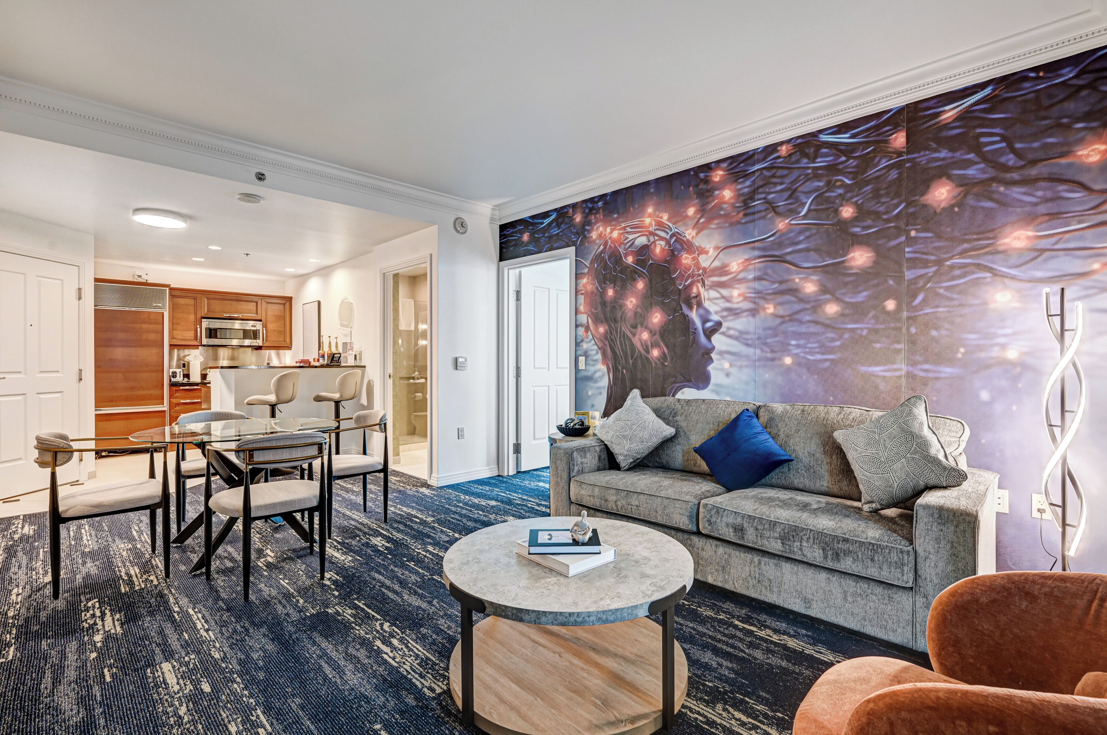 A modern hotel room with a dining area, a gray sofa with multiple pillows, a round coffee table, and a large wall mural.