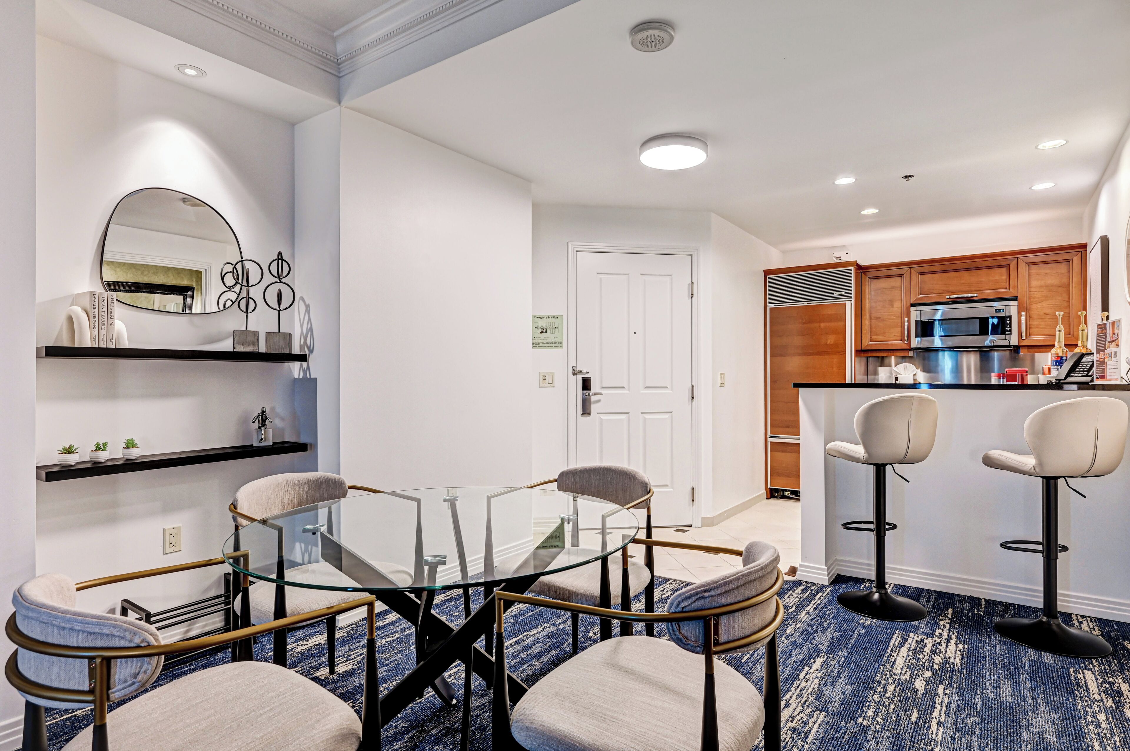 A modern hotel room with a glass dining table, four chairs, a kitchen area with a microwave, and a bar with stools.