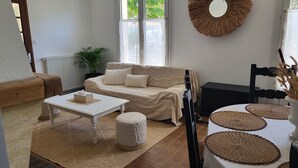 A cozy living room with a sofa, a coffee table, a small ottoman, and a TV. There is a potted plant, a mirror on the wall, and a window with blinds. - Family home in Fouras - enclosed garden near beach and market  (Fouras)