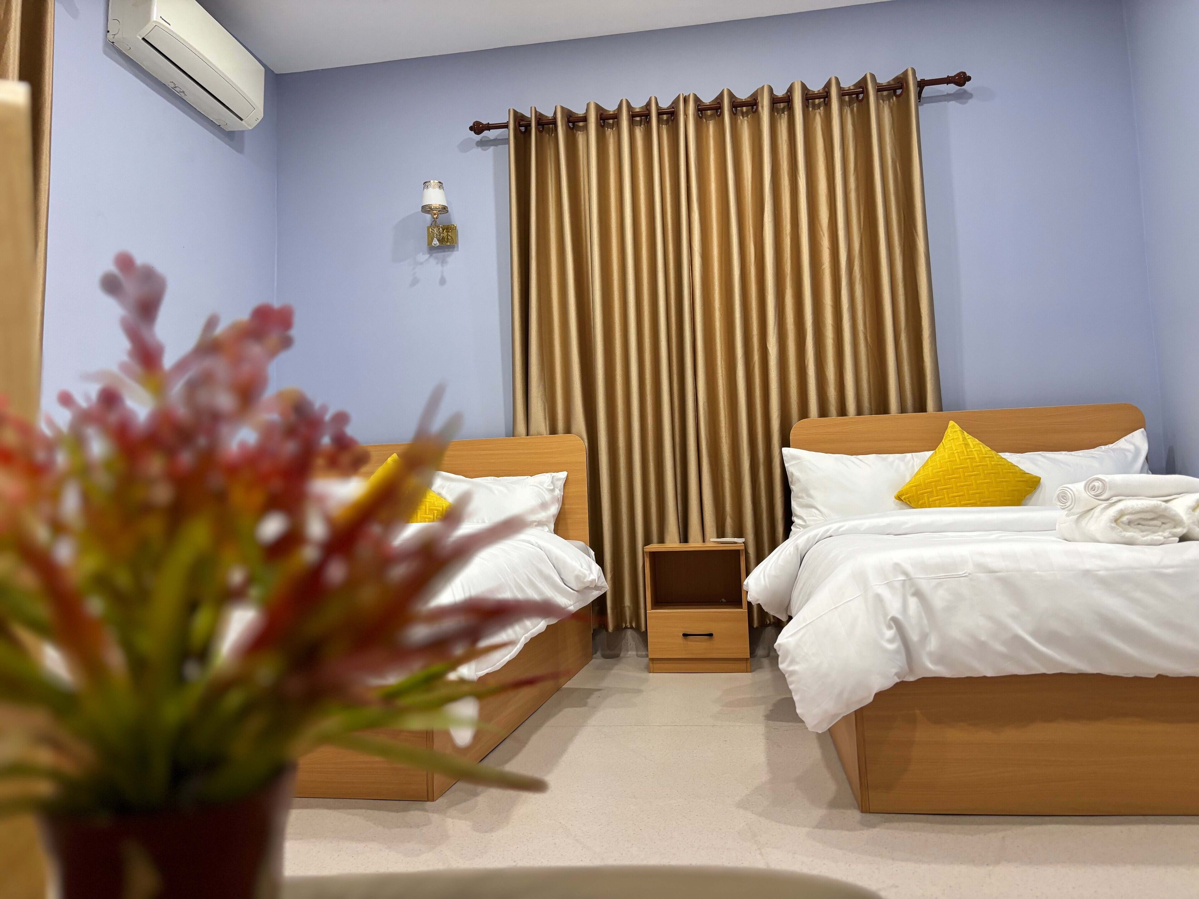 A hotel room with two beds, a potted plant, a nightstand, and a wall-mounted air conditioner.