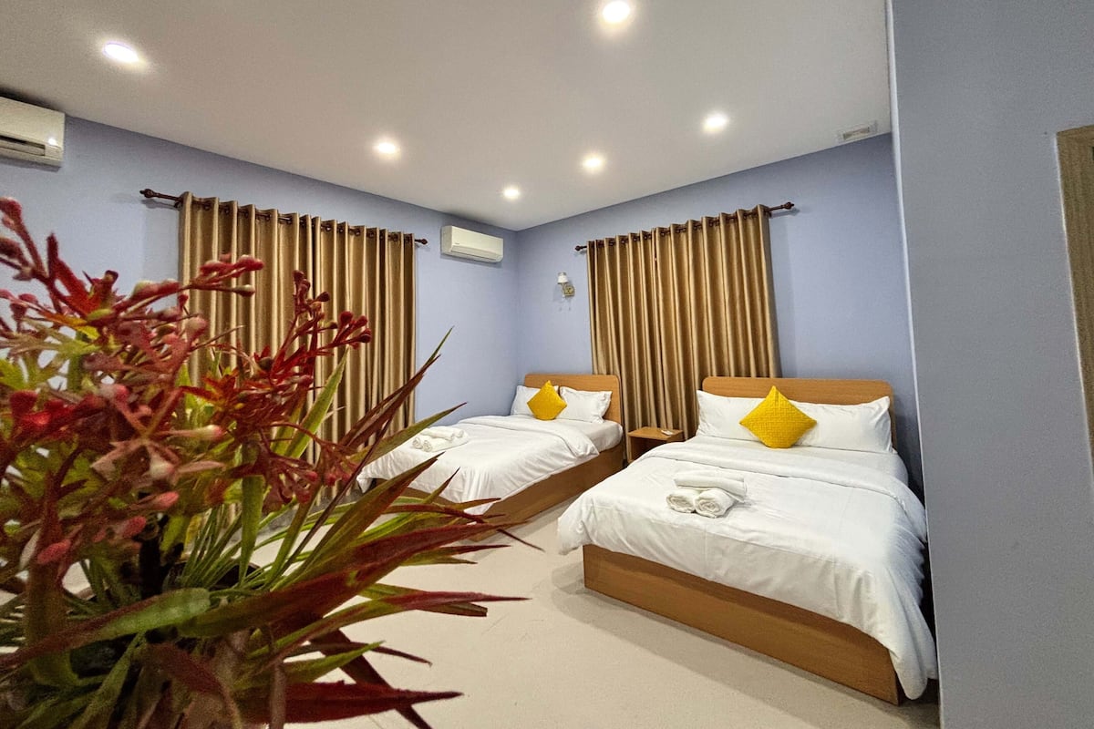 Habitación de hotel con dos camas, cortinas color beige, ropa de cama blanca y una planta en maceta.