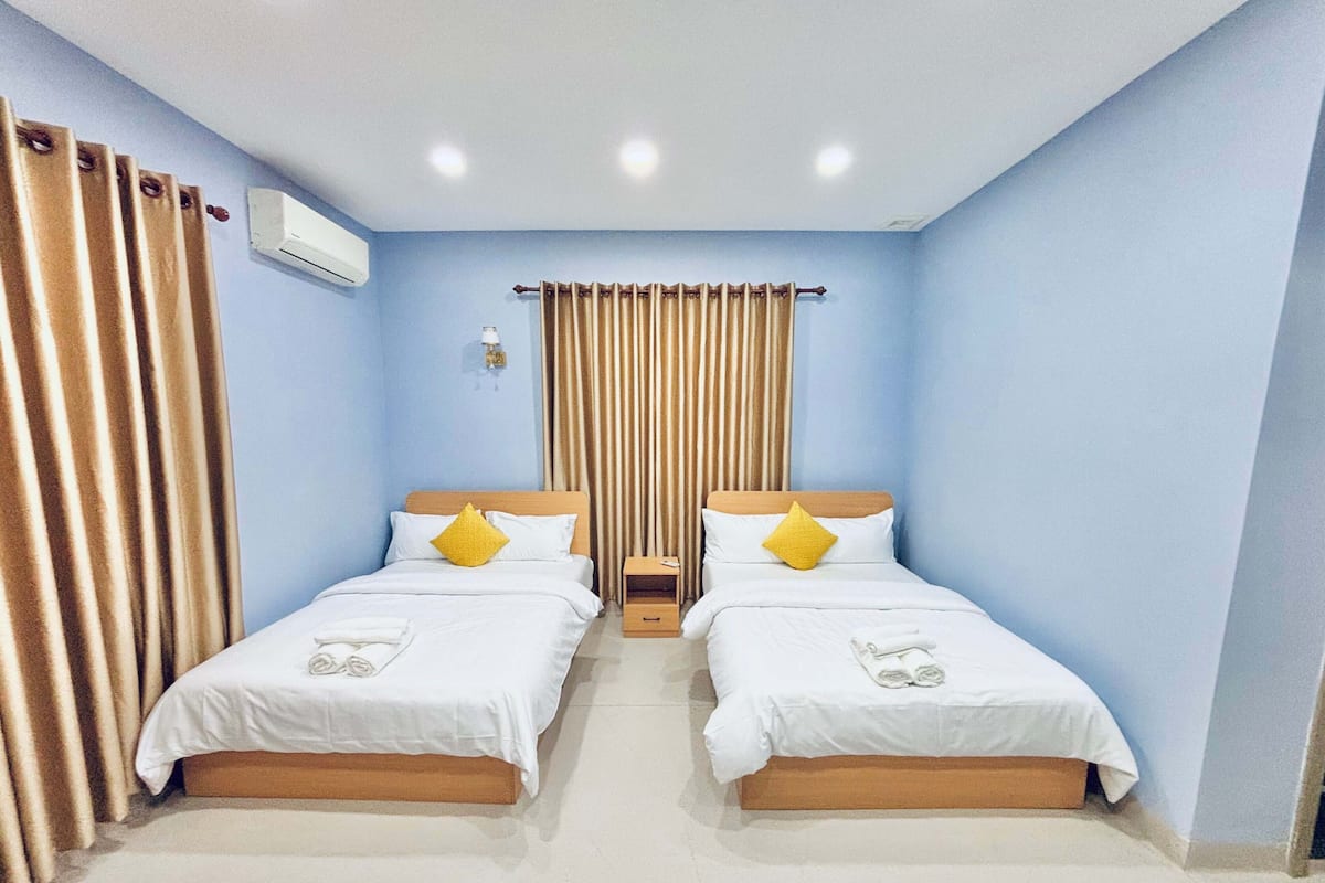 Habitación de hotel con dos camas, cortinas color beige, un aire acondicionado blanco y una mesita con una lámpara.
