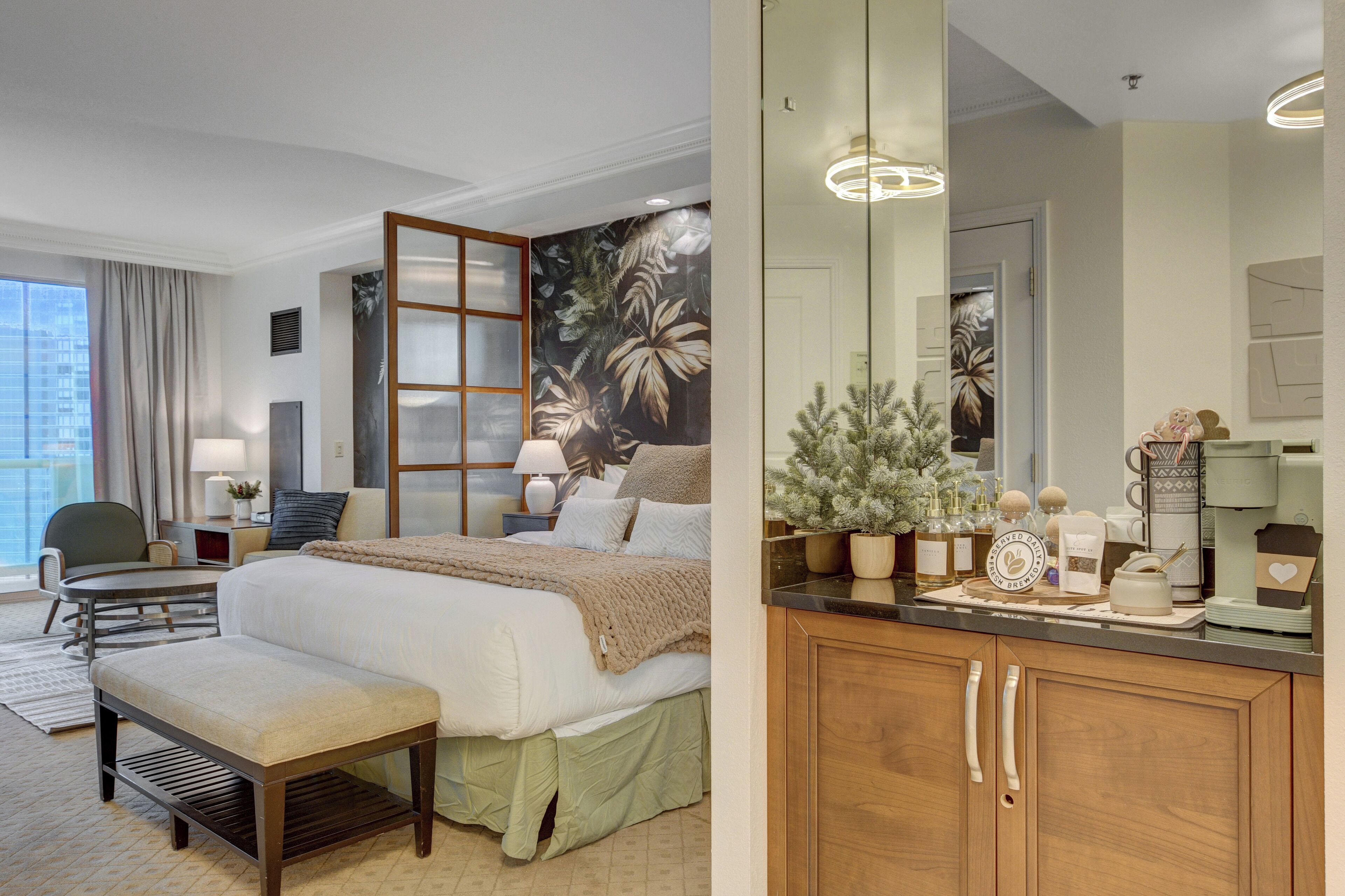 Hotelkamer met een groot bed, een zithoek met een fauteuil en een tafeltje, een houten kast met spiegel en een raam met gordijnen.