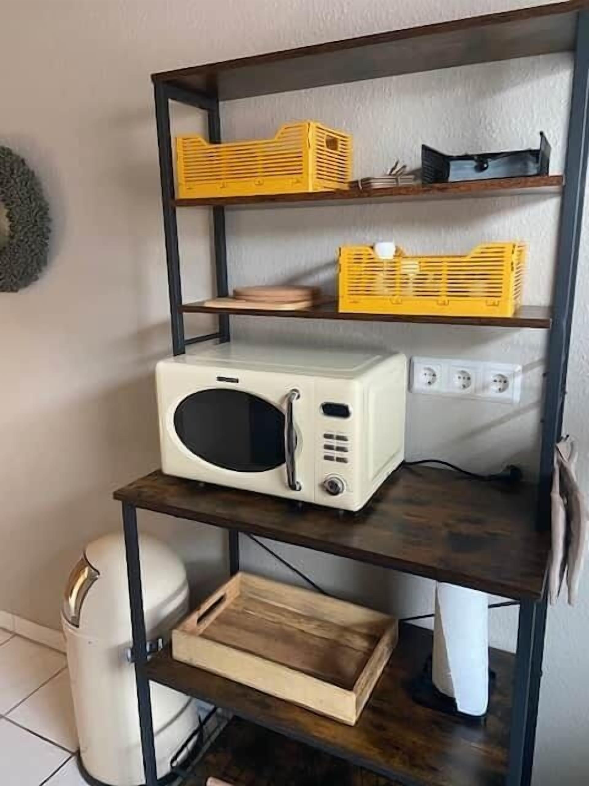 A kitchen area with a microwave on a shelf, a trash can, and a wooden cutting board.