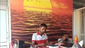 A reception desk with a mural of a sunset over water, a man sitting at the desk, and a bottle of orange juice on the counter. - Sunrise Hotel & Restaurant (Danta Ramgarh)