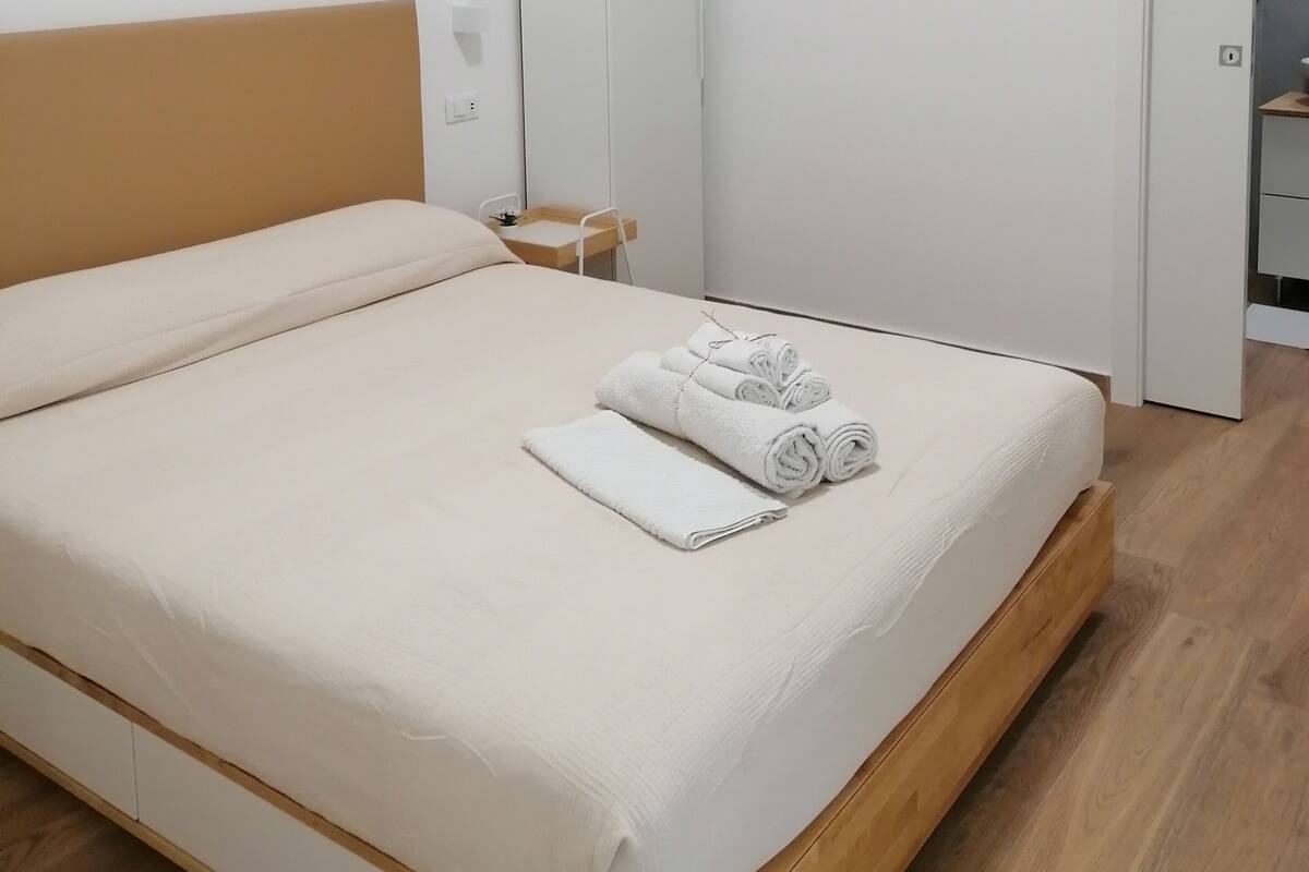 A neatly made bed with white linens and rolled towels, a wooden headboard, a white wardrobe, and a wooden floor.