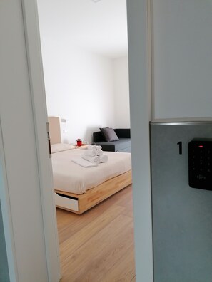 Une chambre d’hôtel moderne avec un plancher en bois, un lit recouvert de draps blancs, une tête de lit noire et une tasse rouge posée sur la table de chevet.