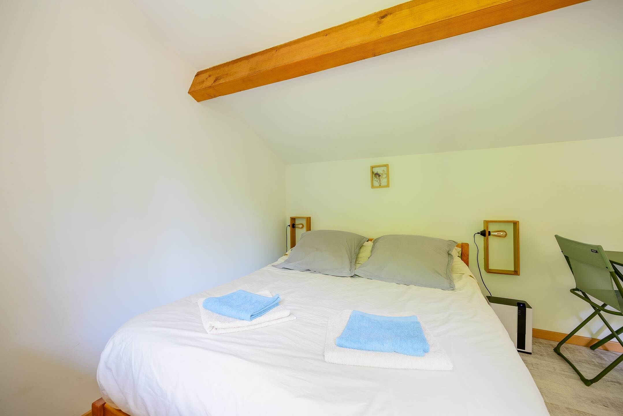 A neatly made bed with white linens and blue towels, a wooden ceiling beam, a small framed picture on the wall, and a folding chair.