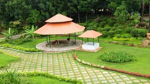 A garden with two umbrella seating areas, a central fire pit, and a paved walkway. - Pragati Resorts (Shankarpalle)