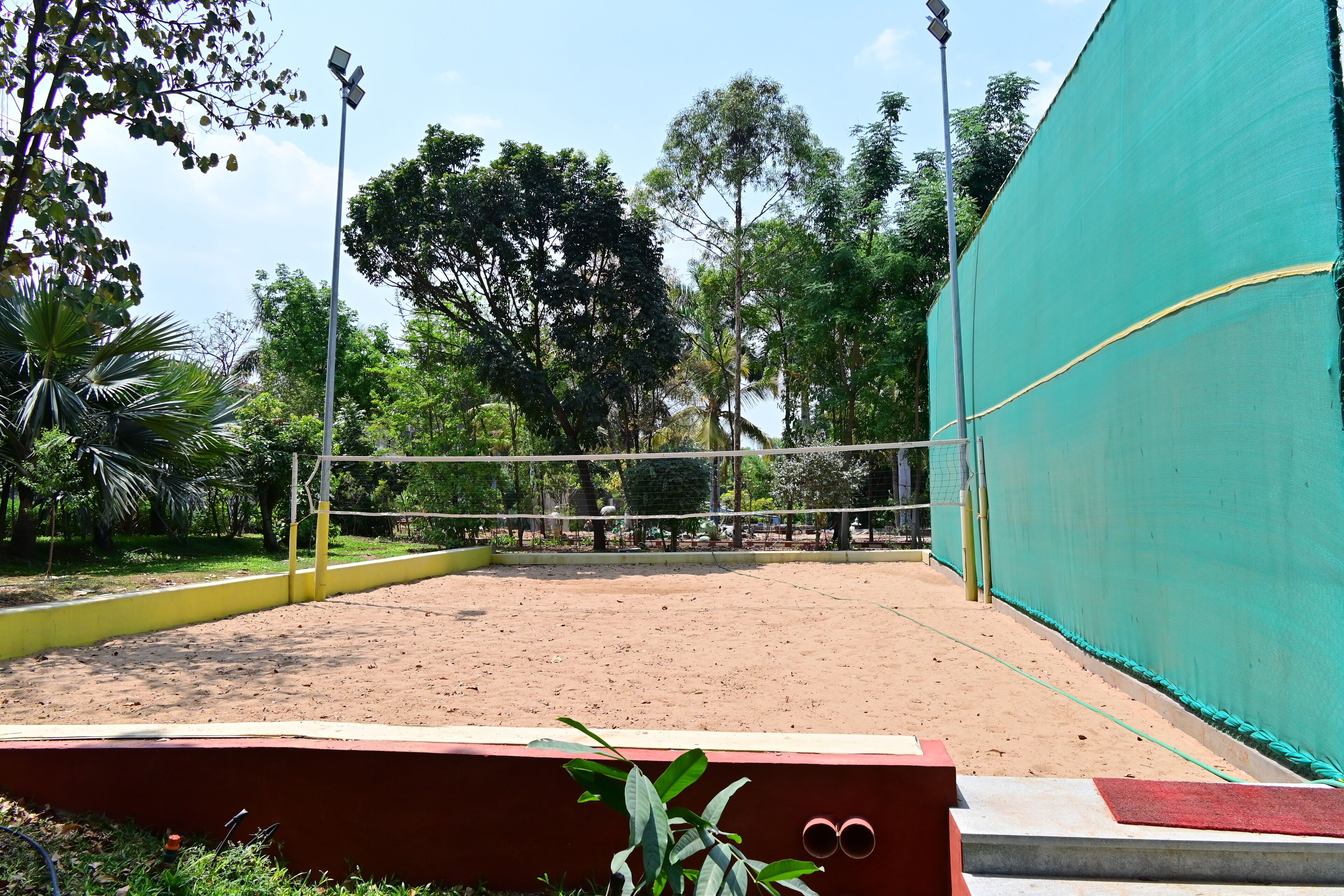 An outdoor volleyball court with a net, surrounded by trees and a green fence.