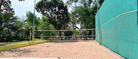An outdoor volleyball court with a net, surrounded by trees and a green fence.