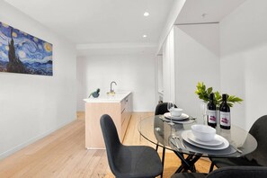 A modern dining area with a glass table, black chairs, a white island with a sink, and a painting on the wall.