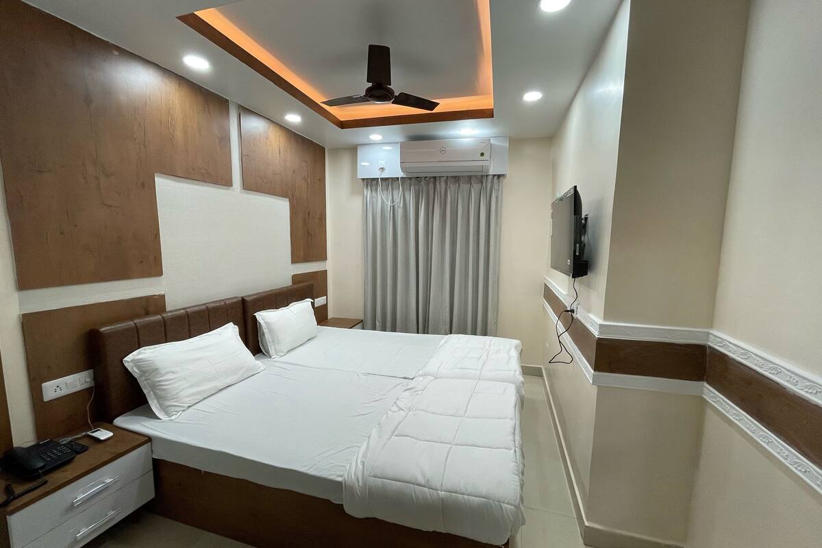 A hotel room with a large bed, a ceiling fan, an air conditioning unit, a television, and a bedside table with a phone and charger.