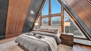 A bedroom with a large bed, two bedside tables, and a window with a view of trees. - Bliss A-Frame, Heated Pool AxeThrow Sundeck Sauna HotTub FirePit (Jim Thorpe)