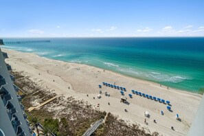 Ein Strand mit einem Pier im Hintergrund, sandigem Ufer und einer Reihe blauer Sonnenschirme.